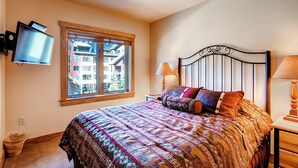 A bedroom with a bed, a TV mounted on the wall, a window with a view of buildings, and a nightstand with a lamp. - Keystone 2BR | Pool Access | Close to the Gondola (Keystone)