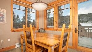 A wooden dining table with four chairs, large windows with a view of trees and a cabin, and a ceiling light fixture. - Gorgeous Ski Escape with Fireplace and Balcony (Keystone)