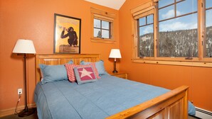 A bedroom with a wooden bed, two bedside lamps, a poster on the wall, and a window with a view of a forest. - Gorgeous Ski Escape with Fireplace and Balcony (Keystone)