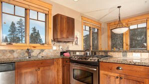 A kitchen with wooden cabinets, granite countertops, a stainless steel stove, and large windows overlooking a forest. - Gorgeous Ski Escape with Fireplace and Balcony (Keystone)