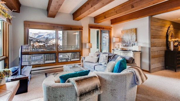 A cozy living room with a sofa, a coffee table, and a TV. There are large windows with a view of mountains. - Condo | 100 Yards from the Slopes | Cozy + Elegant (Crested Butte)