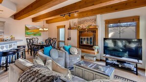 A spacious living room with a high ceiling, wooden beams, a fireplace, a television, and a dining area with a table and chairs. - Condo | 100 Yards from the Slopes | Cozy + Elegant (Crested Butte)