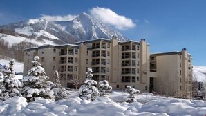 A multi-story hotel with a snow-covered landscape and mountains in the background. - Condo | 100 Yards from the Slopes | Cozy + Elegant (Crested Butte)