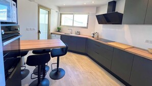 A modern kitchen with dark cabinets, a wooden countertop, and a built-in oven. There are two black bar stools and a window providing natural light.