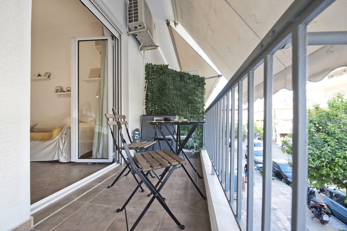 A balcony with a table and chairs, a green wall, and a view of parked cars and greenery.