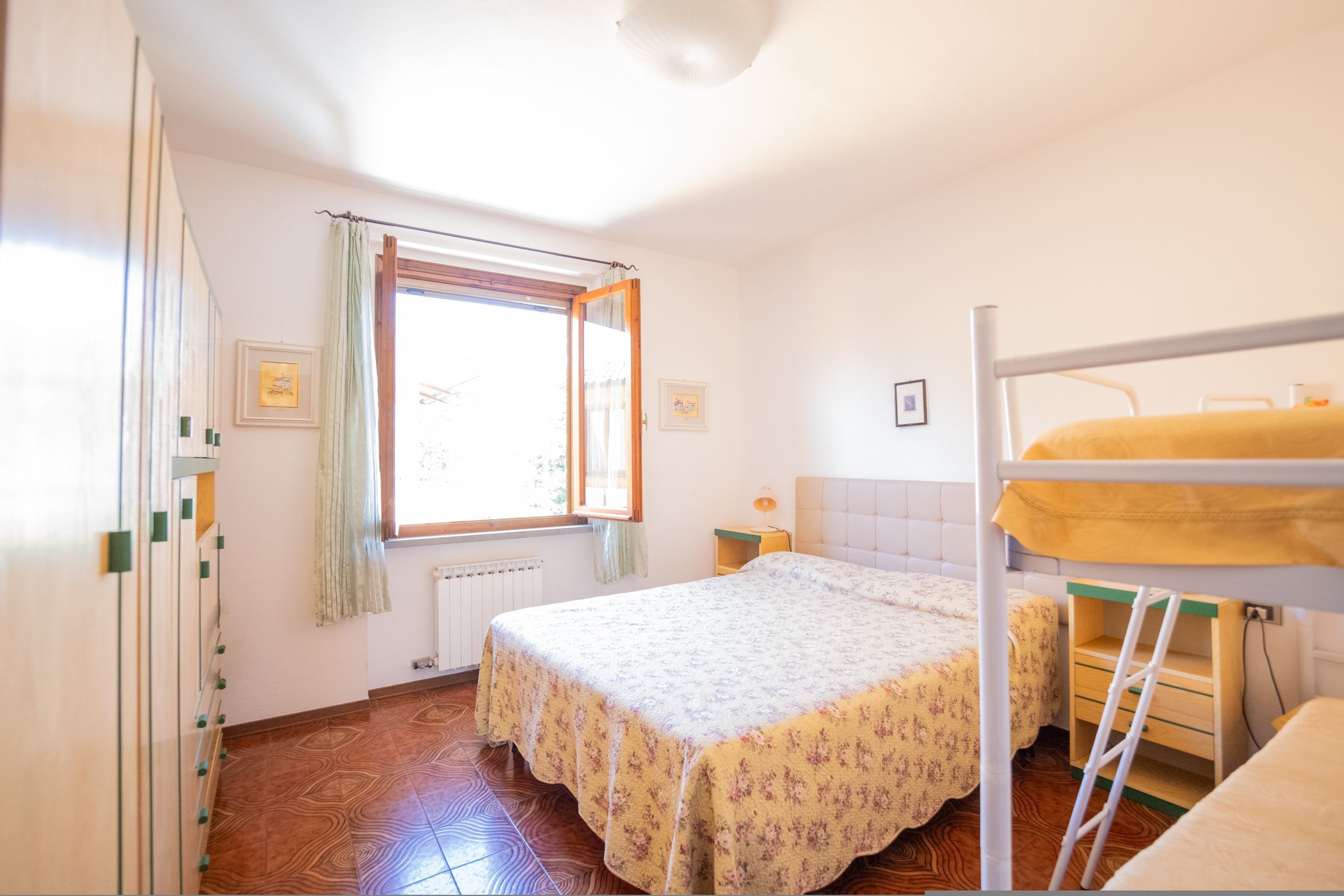 A room with a bunk bed, a radiator, a window with curtains, and a wooden floor.