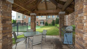 An outdoor patio with a glass table and chairs, a barbecue grill, and a covered area with wooden beams and brick walls.