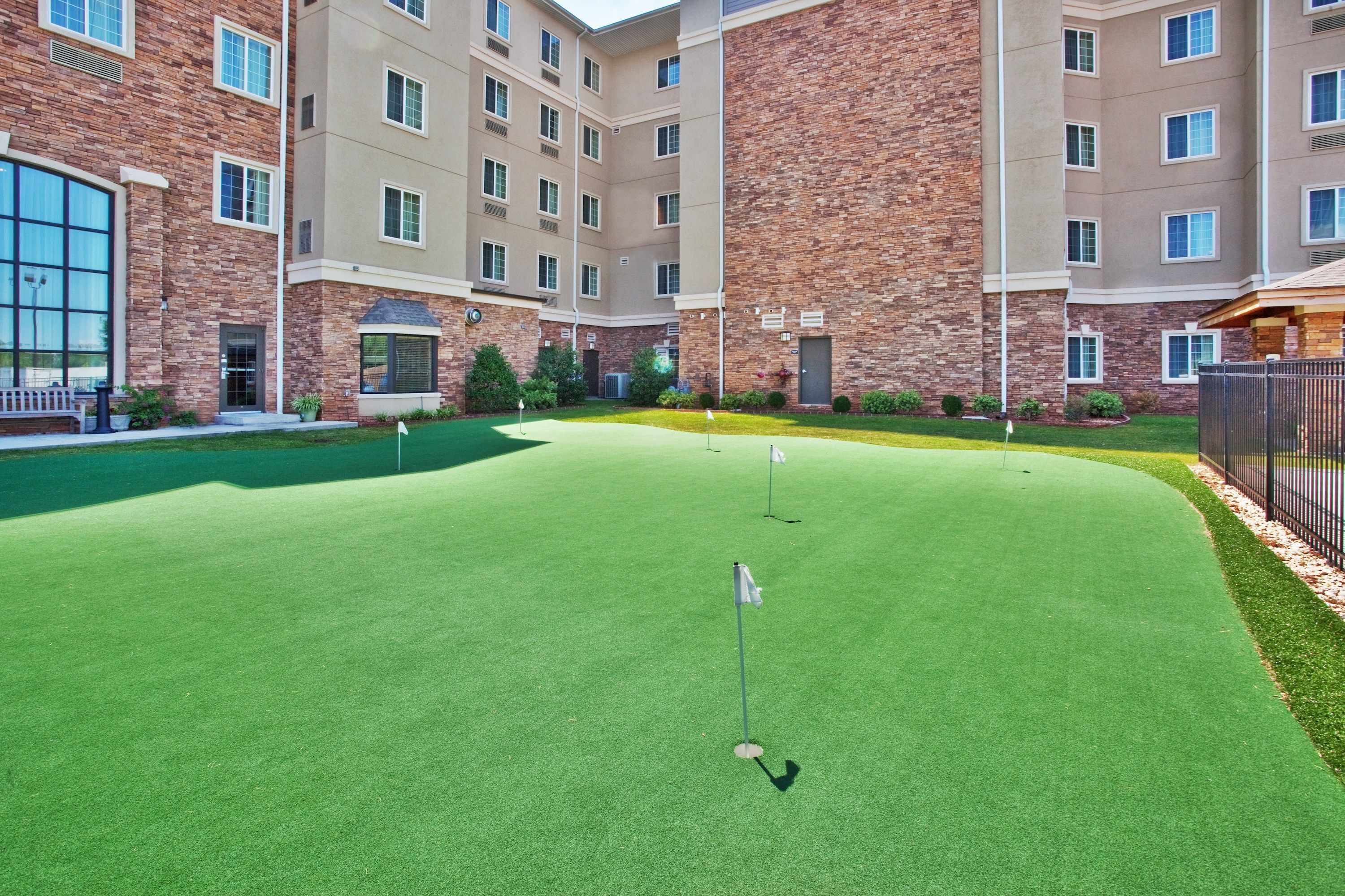 Un parcours de golf doté d’un green d’entraînement, entouré d’un immeuble à plusieurs étages aux murs en brique et de couleur beige, avec de grandes fenêtres et une entrée sécurisée par un portail.