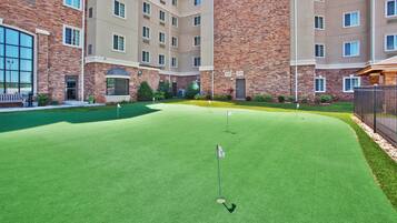 Un parcours de golf doté d’un green d’entraînement, entouré d’un immeuble à plusieurs étages aux murs en brique et de couleur beige, avec de grandes fenêtres et une entrée sécurisée par un portail.