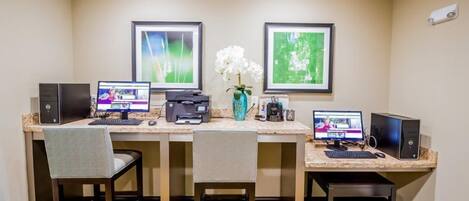 A hotel room with a desk, two computers, a printer, a vase with flowers, and framed pictures on the wall.