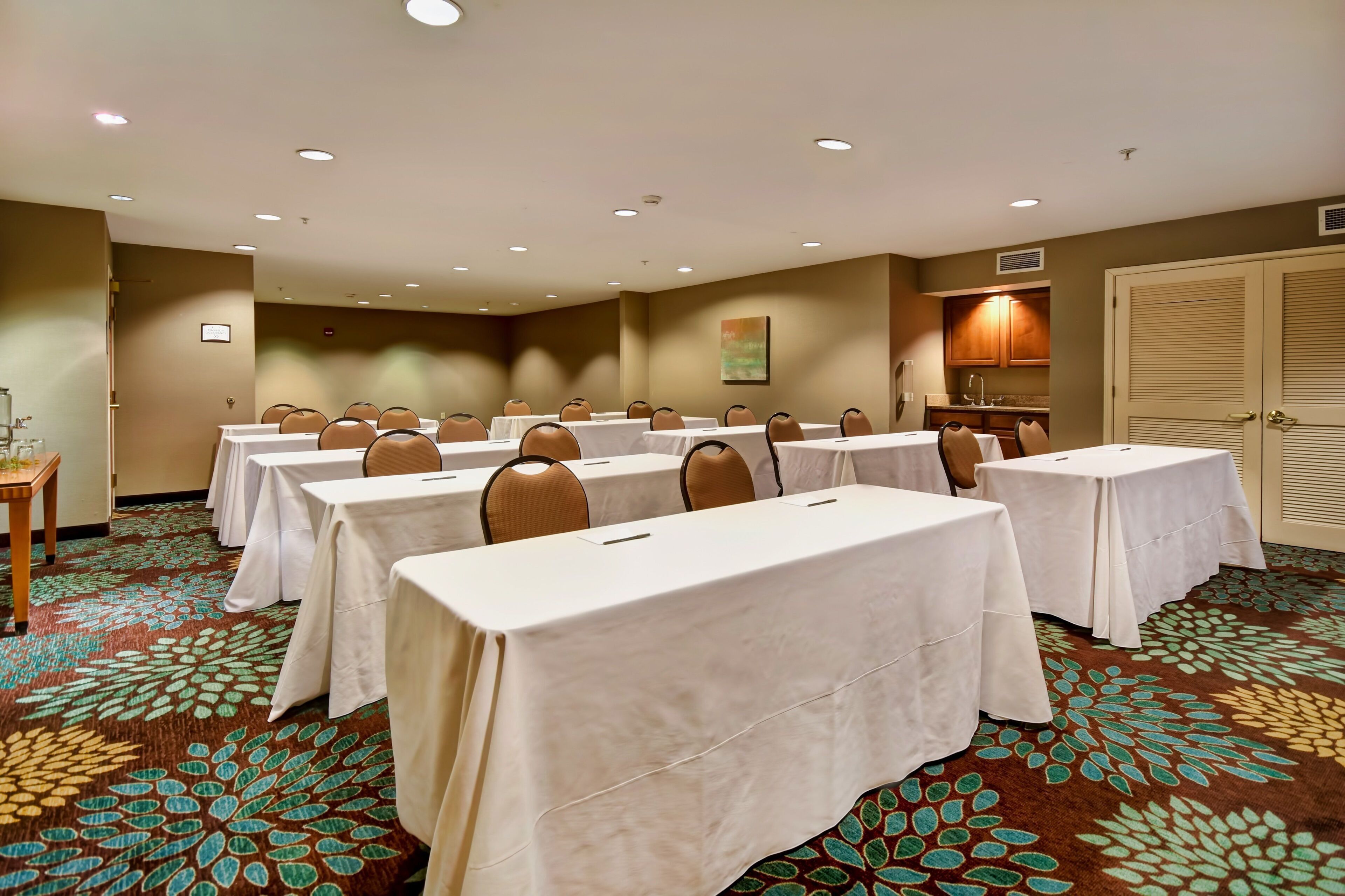 A conference room with multiple tables and chairs, carpeted floor, and a kitchenette area.