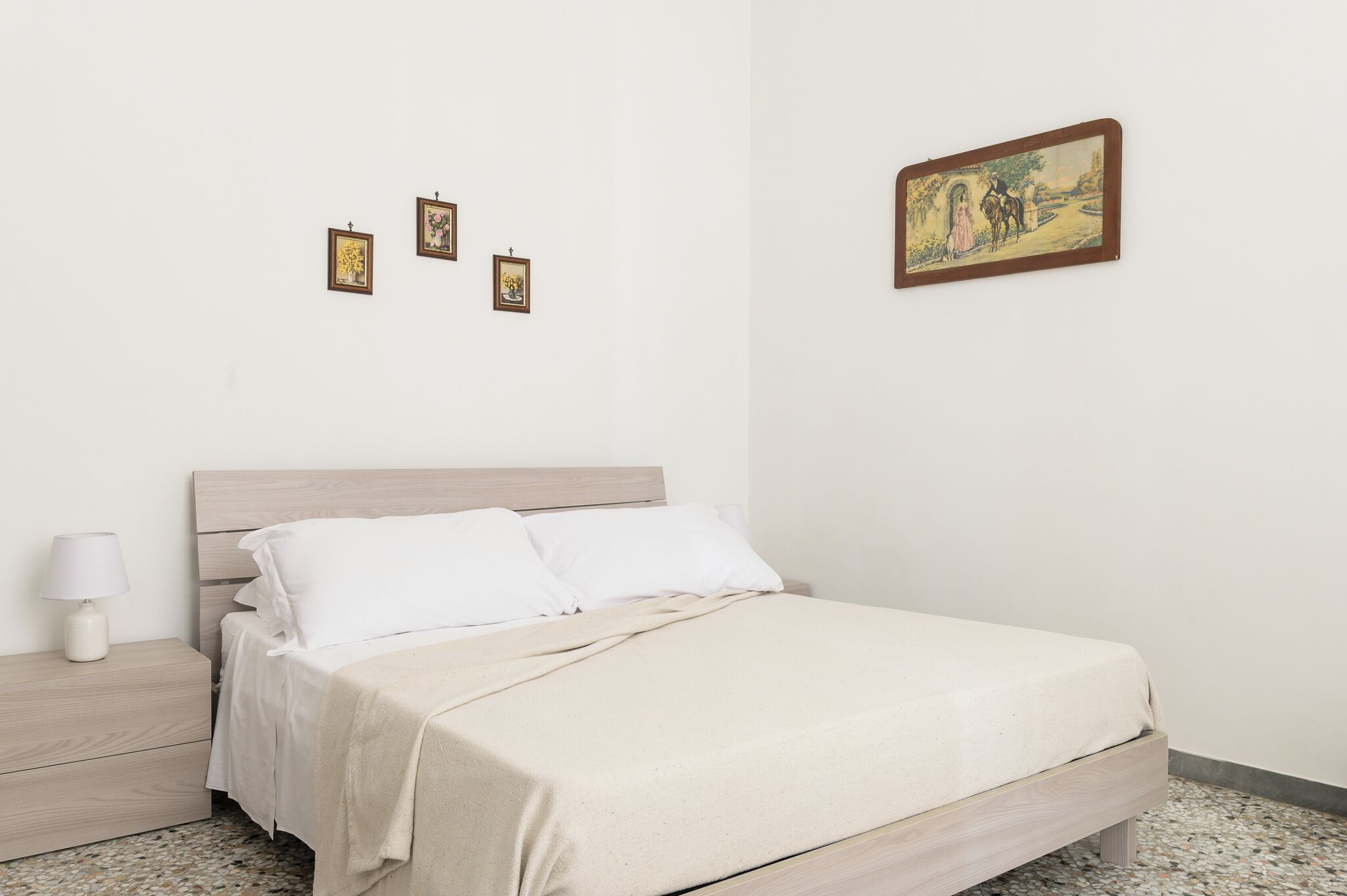 Un lit bien fait, avec du linge de lit blanc, une tête de lit en bois et une couverture beige. Trois petites photos encadrées sont accrochées au mur, au-dessus du lit.
