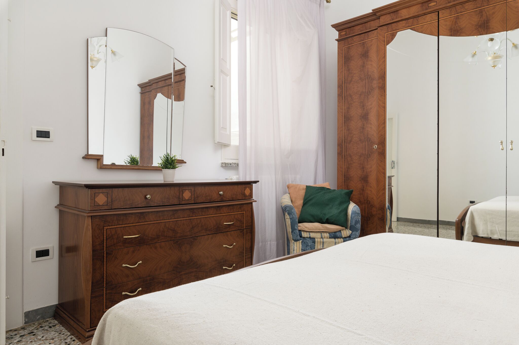 A bedroom with a wooden dresser, a bed, a mirror, and a window with curtains.
