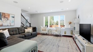 A modern living room with a sectional sofa, a flat-screen TV, and a large window with blinds. - Spacious 4BR-4BA House for Large Families & Group in Stonebridge (Saskatoon)