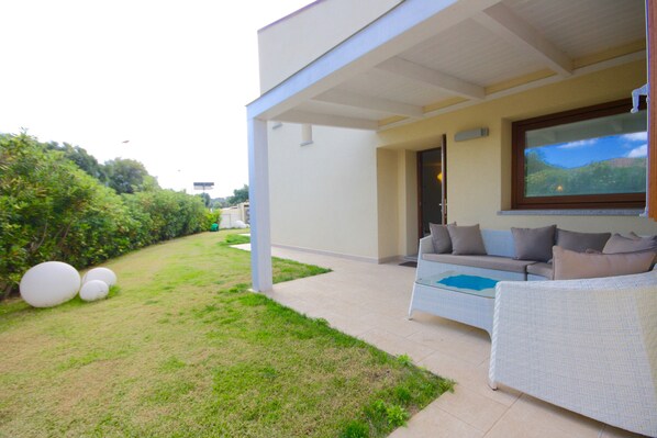 A covered outdoor seating area with a sofa and cushions, a large window, and a grassy lawn with spherical decorations.