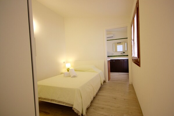 A narrow hallway leading to a bedroom with a bed, a bedside lamp, and a bathroom visible in the background.