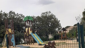 A playground with a slide and climbing structures, surrounded by a fence and various plants. - Maison et Loisirs (Calvisson)