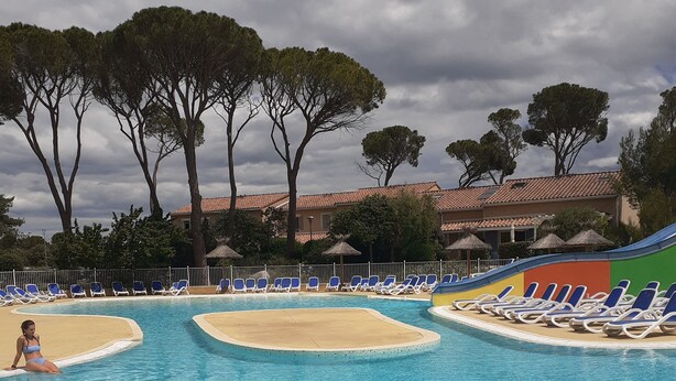A swimming pool with clear blue water, surrounded by lounge chairs and umbrellas, with trees and a building in the background. - Maison et Loisirs (Calvisson)