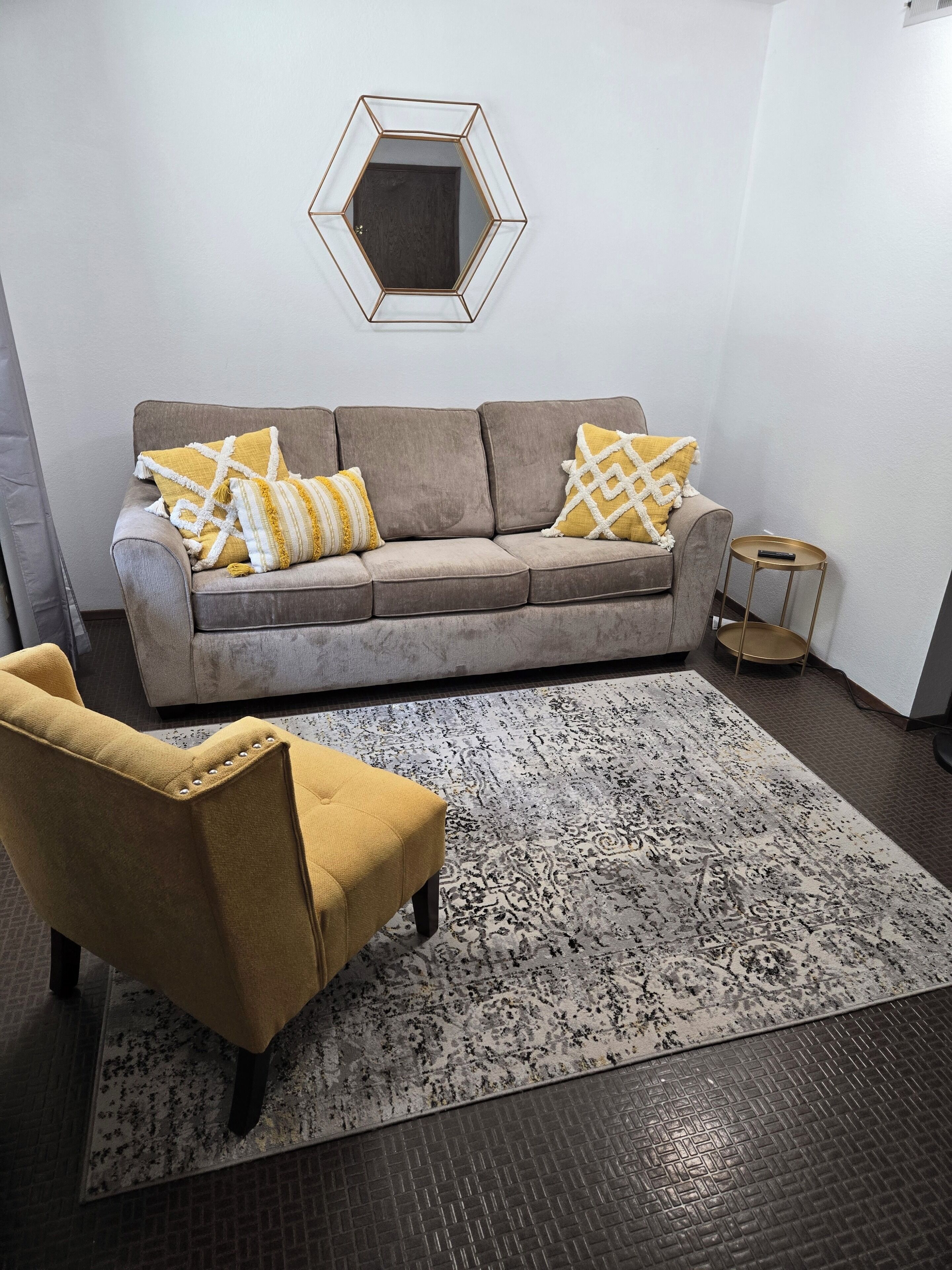 Un salon avec un canapé gris, deux coussins jaunes, un fauteuil de couleur moutarde, un tapis à motifs et un miroir hexagonal accroché au mur.