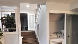Ein Waschraum mit einer Waschmaschine und einem Trockner, einer Treppe zu einer oberen Ebene sowie einer Zimmerpflanze in der Ecke.