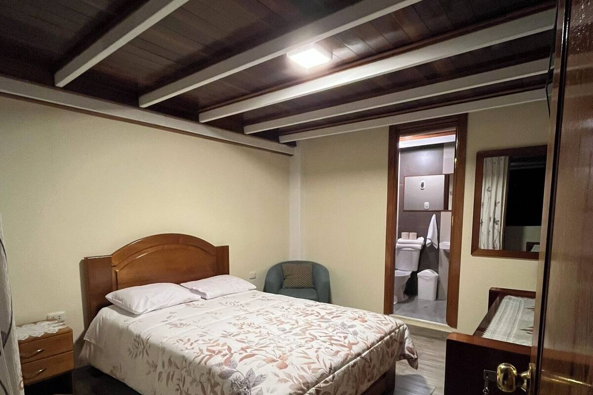 A bedroom with a wooden bed, a nightstand, a chair, a wardrobe, and a door leading to a bathroom.