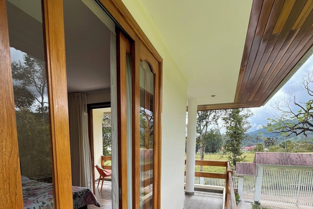 A balcony with wooden railings, a view of a garden, and a bedroom with a bed and curtains.