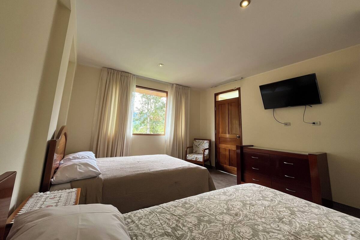 A hotel room with two beds, a TV mounted on the wall, a window with curtains, and a wooden door.
