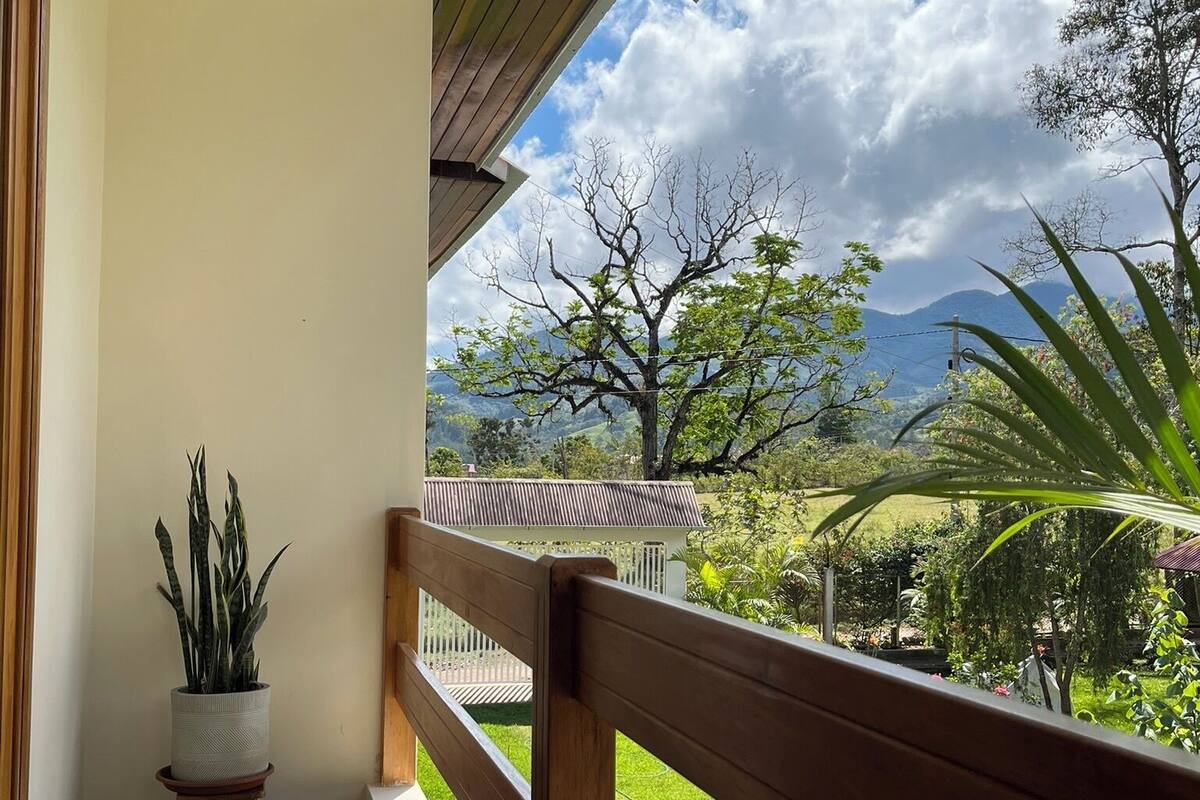 A balcony with a wooden railing, a potted plant, and a view of a grassy area with trees and mountains in the background.