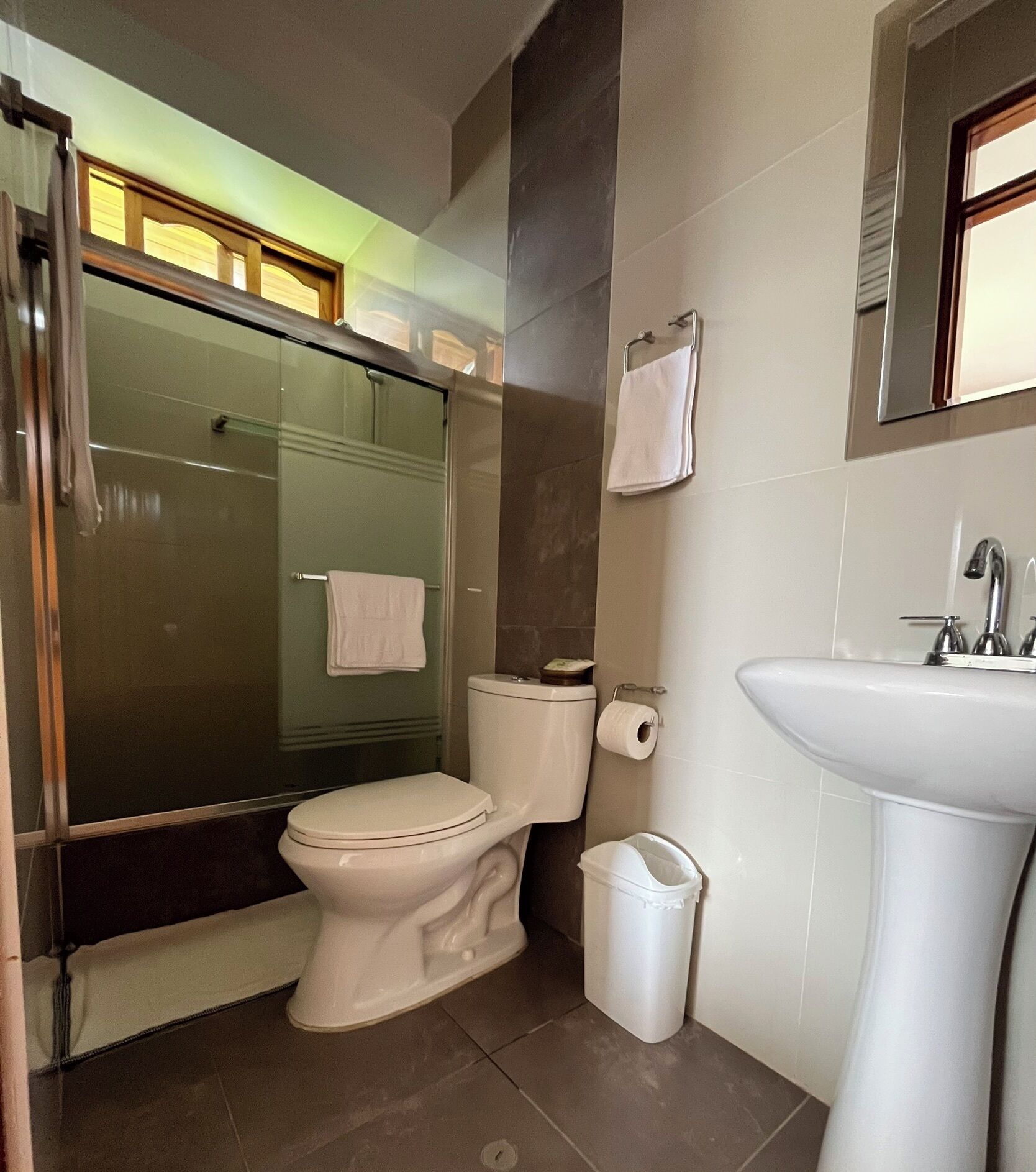 Un baño con inodoro, lavamanos, cabina de ducha de vidrio y un cubo de basura.
