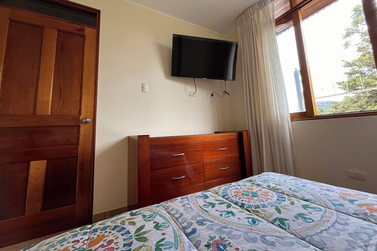 A bedroom with a wooden wardrobe, a bed with a patterned bedspread, a television mounted on the wall, and a window with curtains.