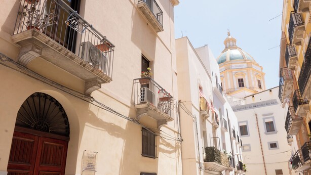 A narrow street with buildings featuring balconies, a prominent dome in the background, and a red door on the left. - Room 2 A (Trapani)
