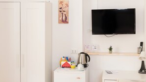 Ein kleiner, sauberer Raum mit einem weißen Schrank, einem weißen Schreibtisch mit Stuhl, einem kleinen Kühlschrank, einem flachbildschirmfernseher an der Wand und einem kleinen Regal mit Pflanze und Wasserkocher.