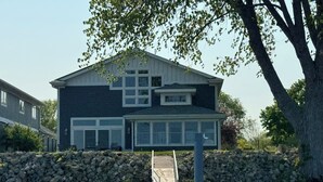 A two-story house with a stone wall in front, a dock extending into a body of water, and a tree to the right.