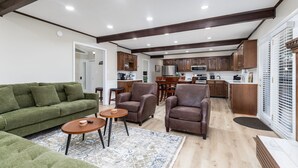 A spacious living area with a green sofa, two armchairs, a coffee table, and a patterned rug. A kitchen with wooden cabinets and a dining area with a table and chairs is visible in the background. - Becca's Place - Near LeConte + Ripken | Pet-Friendly w/ Hot Tub & Games (Pigeon Forge)