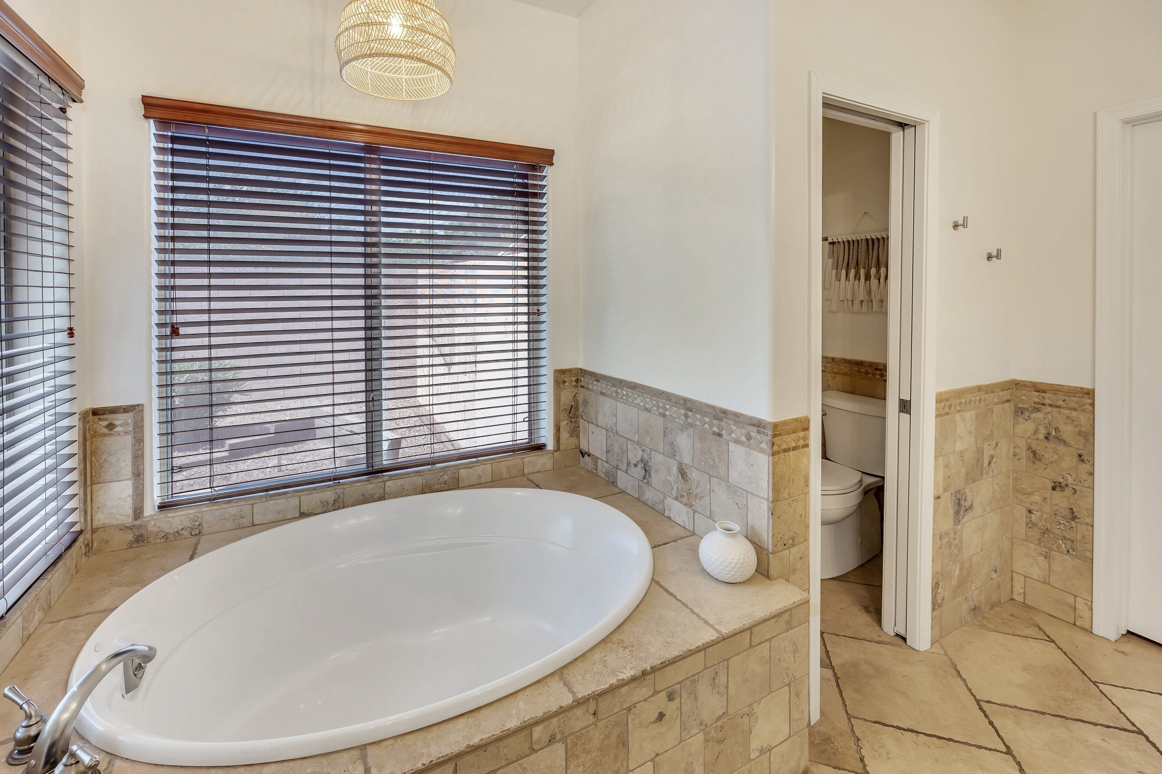 Een badkamer met een groot, ovaal bad, een raam met zonwering en een toilet dat door een open deur zichtbaar is.