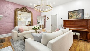 A living room with a pink brick wall, a white sofa set, a wooden piano, and a large mirror. - Luxury Rose Manor - Perfect for Bachelorettes! (Columbus)