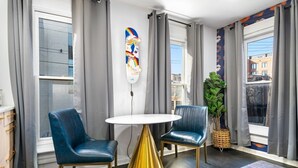 Una habitación moderna con una mesa redonda, dos sillas azules, un adorno en la pared y vistas a edificios a través de amplias ventanas.