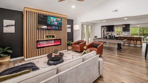 Uma sala de estar moderna com uma TV grande, assentos confortáveis, mesa de bilhar e área de cozinha visível através de uma porta aberta.