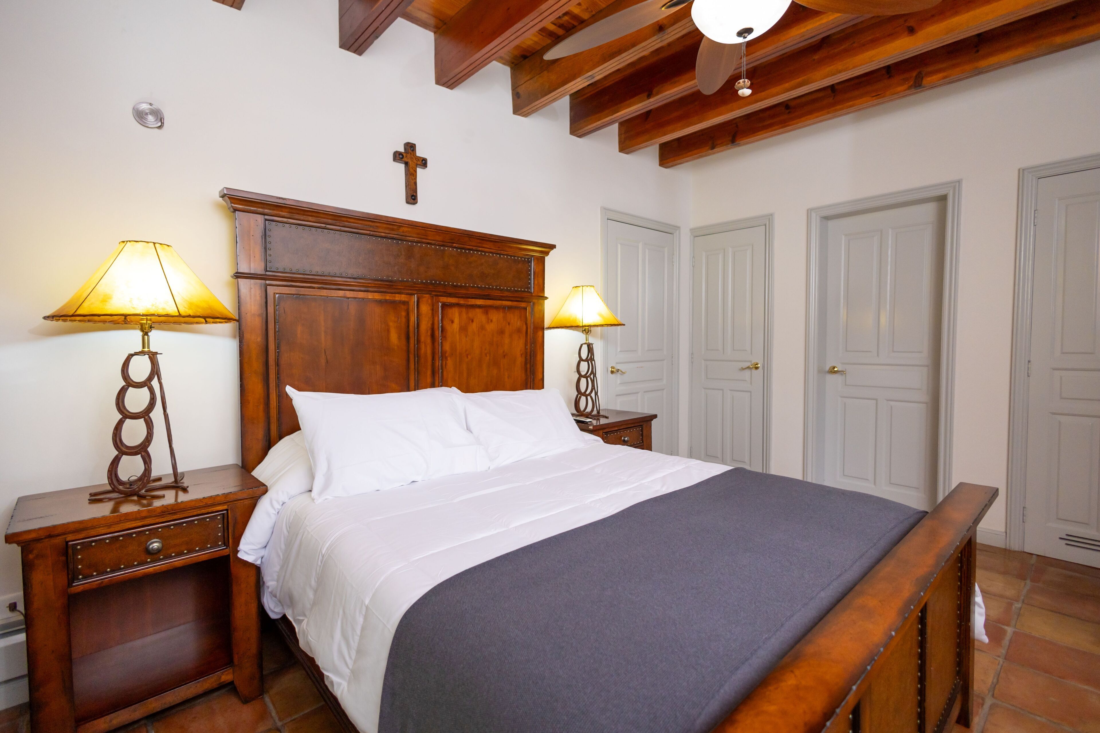 Une chambre à coucher avec un lit en bois, deux tables de chevet avec des lampes, une croix au mur et des portes blanches.