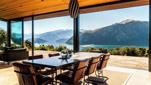 Ein moderner Essbereich mit großem Tisch und Stühlen, große Fenster mit Blick auf Berge und einen See.