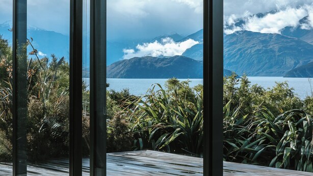 Blick durch ein Fenster auf ein Holzdeck, mit Blick auf einen See und Berge mit Wolken.