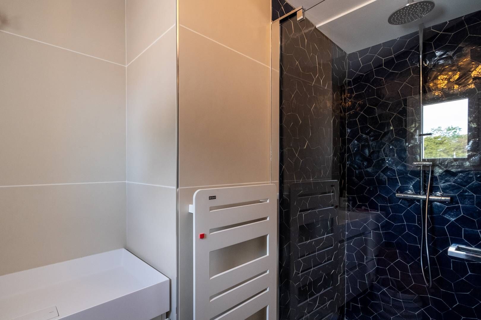 Un baño moderno con un banco blanco, cabina de ducha de vidrio y pared de mosaico negro.
