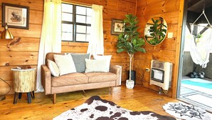 Un intérieur de chalet chaleureux avec un plancher en bois, un canapé, un tapis en peau de vache, une cheminée et une grande fenêtre avec des rideaux.