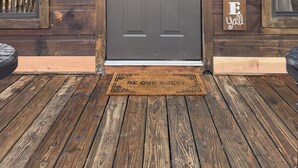 Une cabane en bois avec une porte d’entrée de couleur gris foncé, une pancarte de bienvenue et un paillasson.