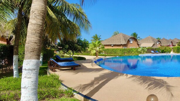 Ein Poolbereich eines Resorts mit Liegestühlen, einem zentralen Pool und Gebäuden mit Reetdächern.