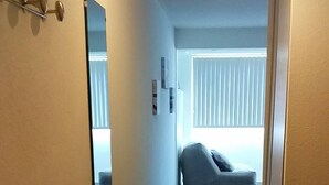 Ein schmaler Flur mit einer grauen Couch im Hintergrund, einem Spiegel links und einer kleinen weißen Regalwand.