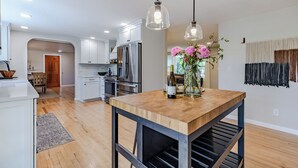 Une cuisine moderne avec des meubles blancs, des appareils en acier inoxydable et une table à manger en bois, surmontée de luminaires suspendus.