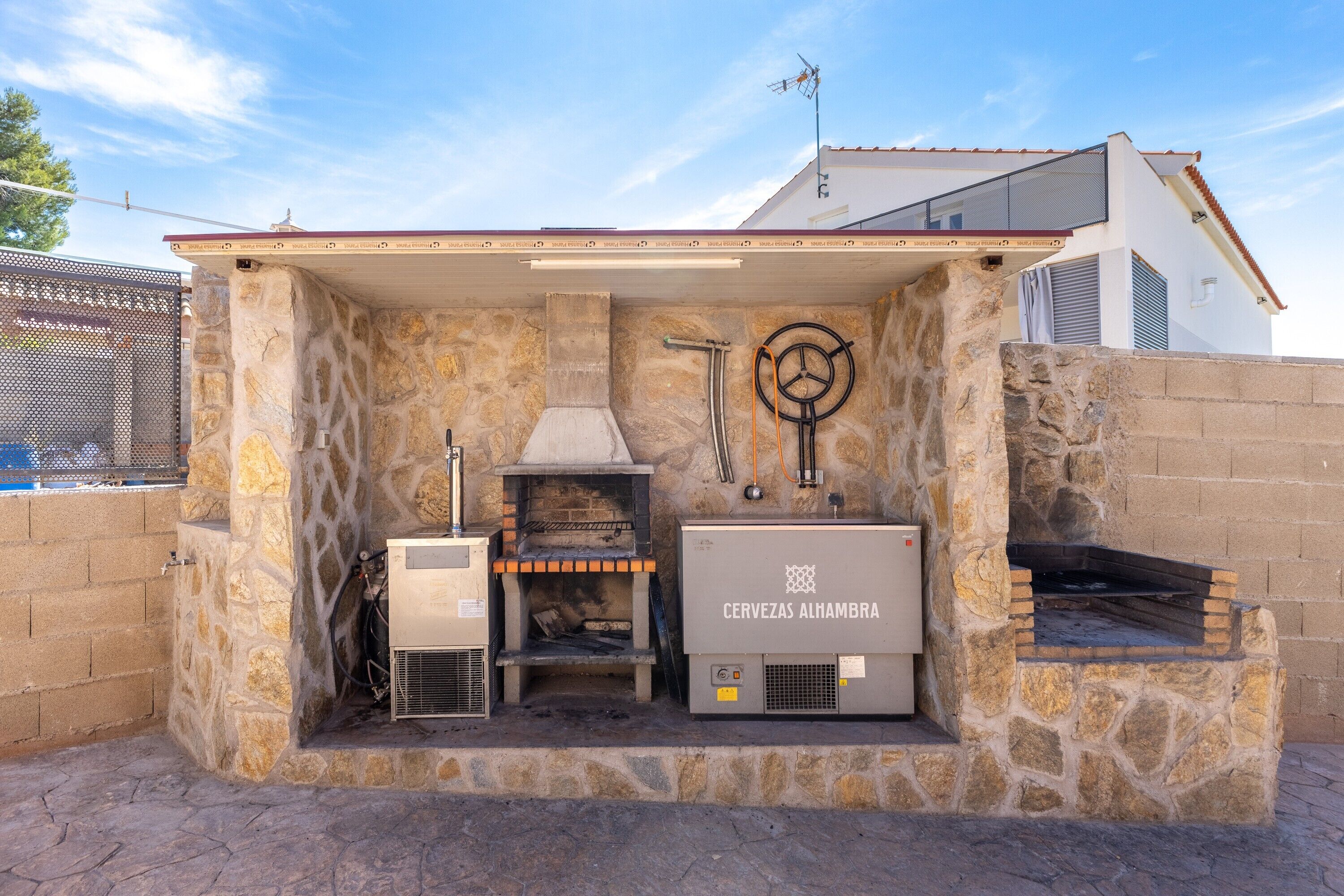 An outdoor kitchen area with a stone wall, a pizza oven, and a beer fridge labeled 'CERVEZAS ALHAMBRA'.