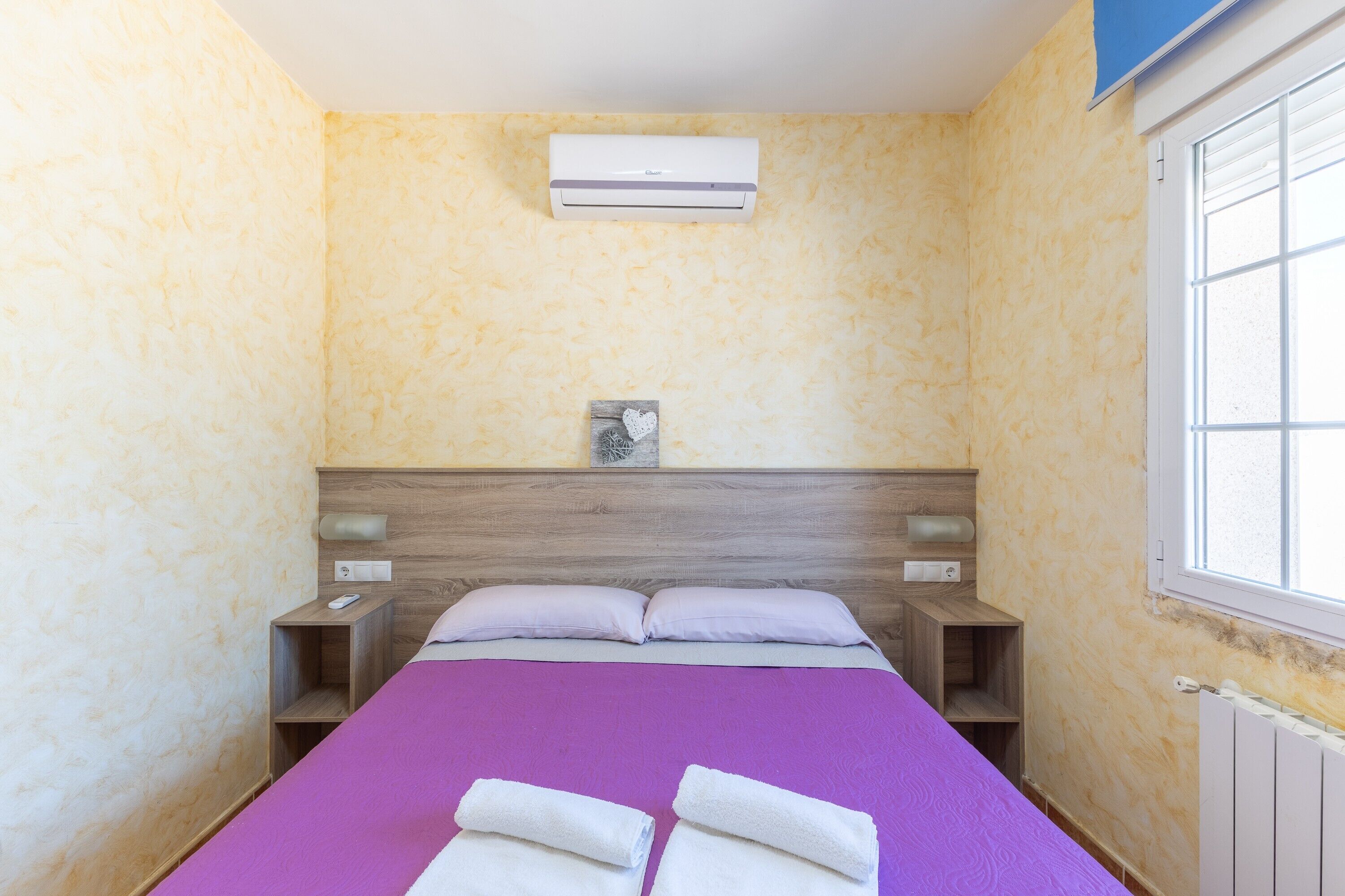 Una habitación de hotel con cama, mesita de noche, aire acondicionado instalado en la pared y ventana.