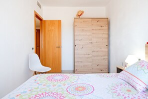 A bedroom with a bed, a wooden wardrobe, a white chair, and a nightstand with a lamp.